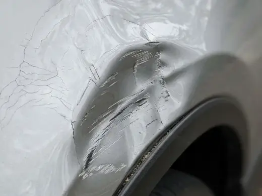 A close-up detail shot of cracked silver paint and creased metal on the car fender.
